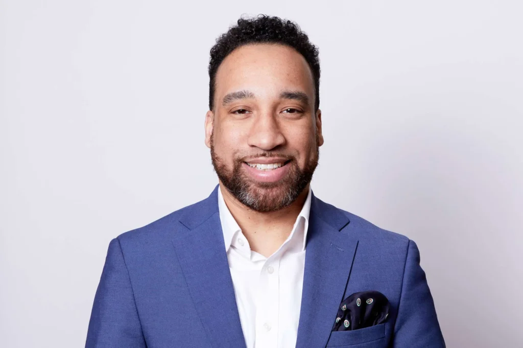 A cheerful man with a friendly smile is dressed in a crisp white shirt and blue suit with a patterned pocket square. His short curly hair and well-groomed beard add to the professional yet approachable look of the portrait set against a soft, neutral background.