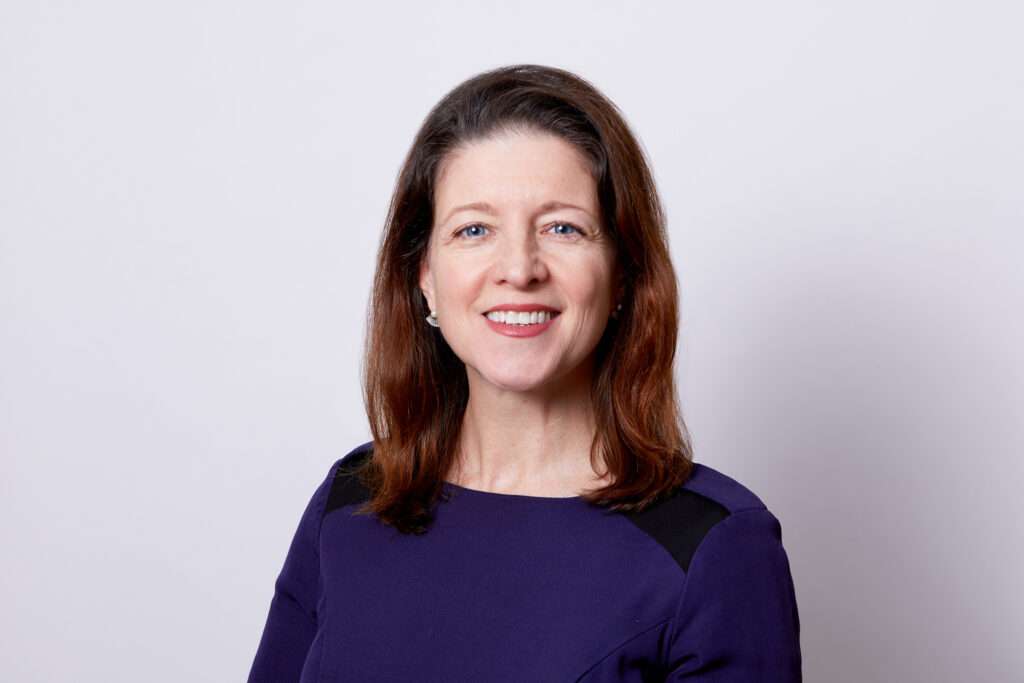 A portrait of a woman with long chestnut hair and blue eyes, smiling warmly. She wears a stylish dark purple dress with black shoulder accents, complemented by subtle earrings. The image conveys a professional and welcoming demeanor against a bright, unobtrusive background.