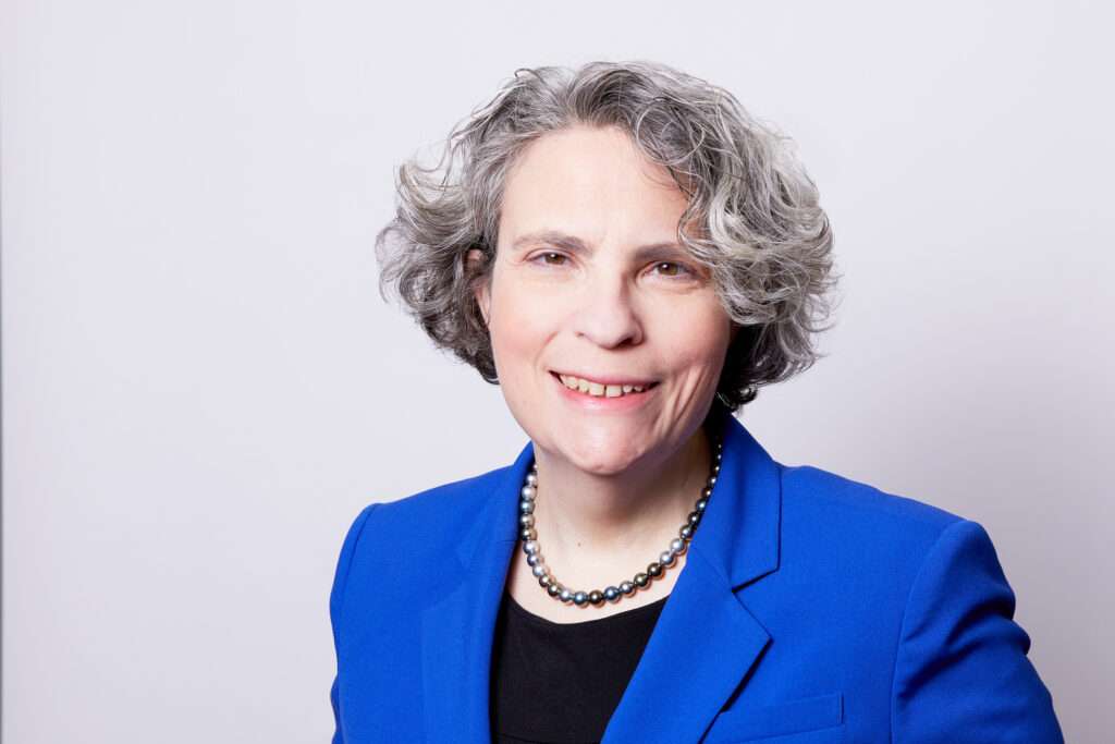A professional portrait of a woman with shoulder-length curly grey hair, featuring a gentle smile. She's dressed in a bold blue blazer over a black top and accessorized with a strand of pearls, projecting a look of confidence and approachability against a soft white background.