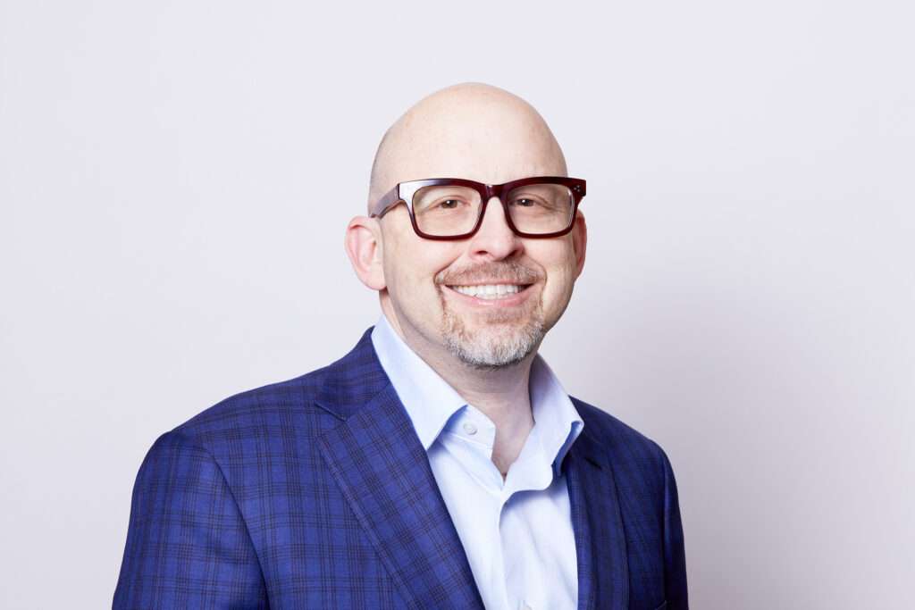 A smiling bald man with glasses is dressed in a blue checked blazer over a light blue shirt. The backdrop is a clean, neutral color, providing a bright and professional appearance to the portrait. His friendly demeanor is conveyed through his relaxed pose and pleasant smile.