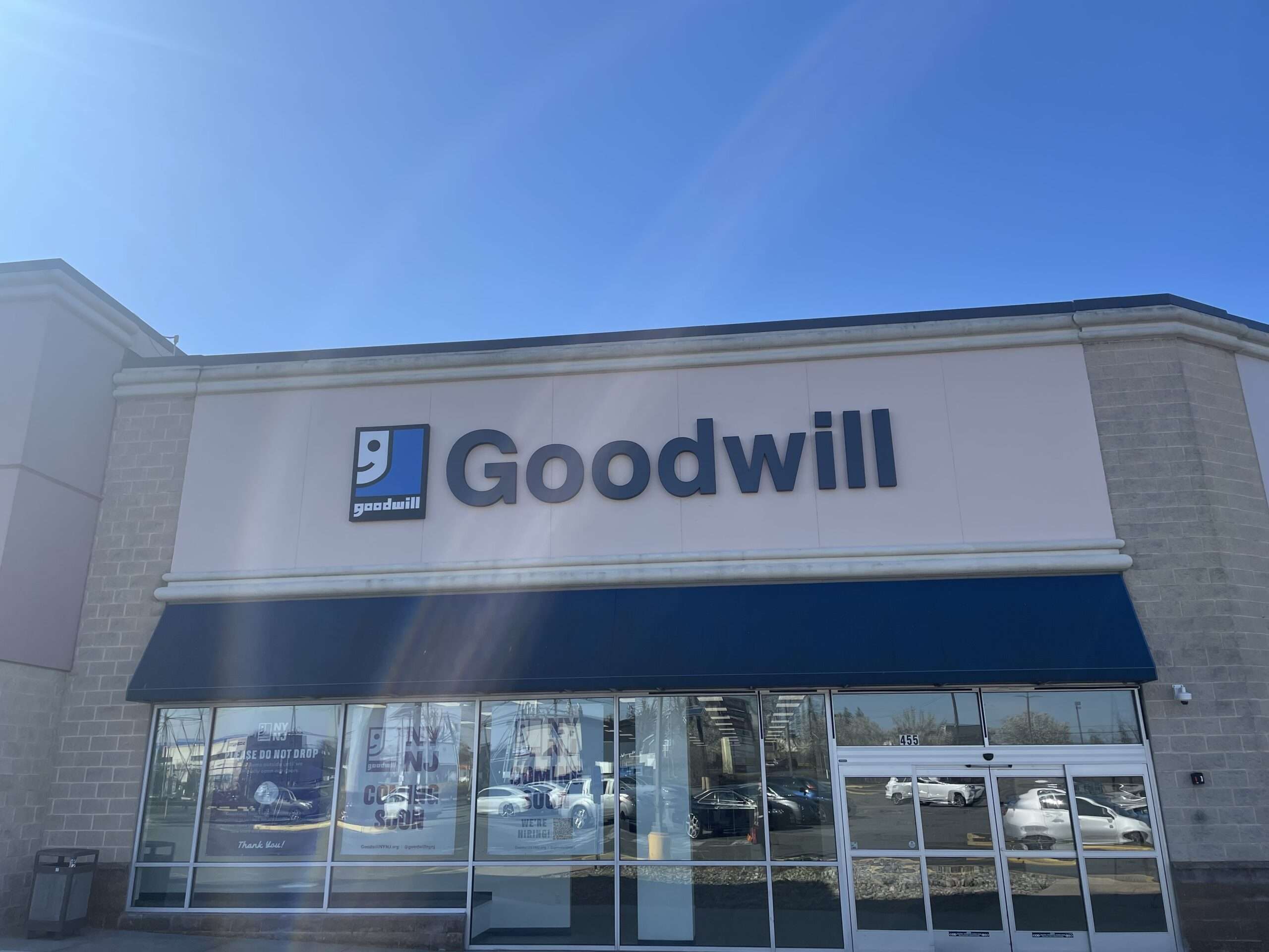 Exterior view of a Goodwill retail store on a sunny day, with the Goodwill logo prominently displayed above the entrance, reflecting a clear blue sky in its large front windows.
