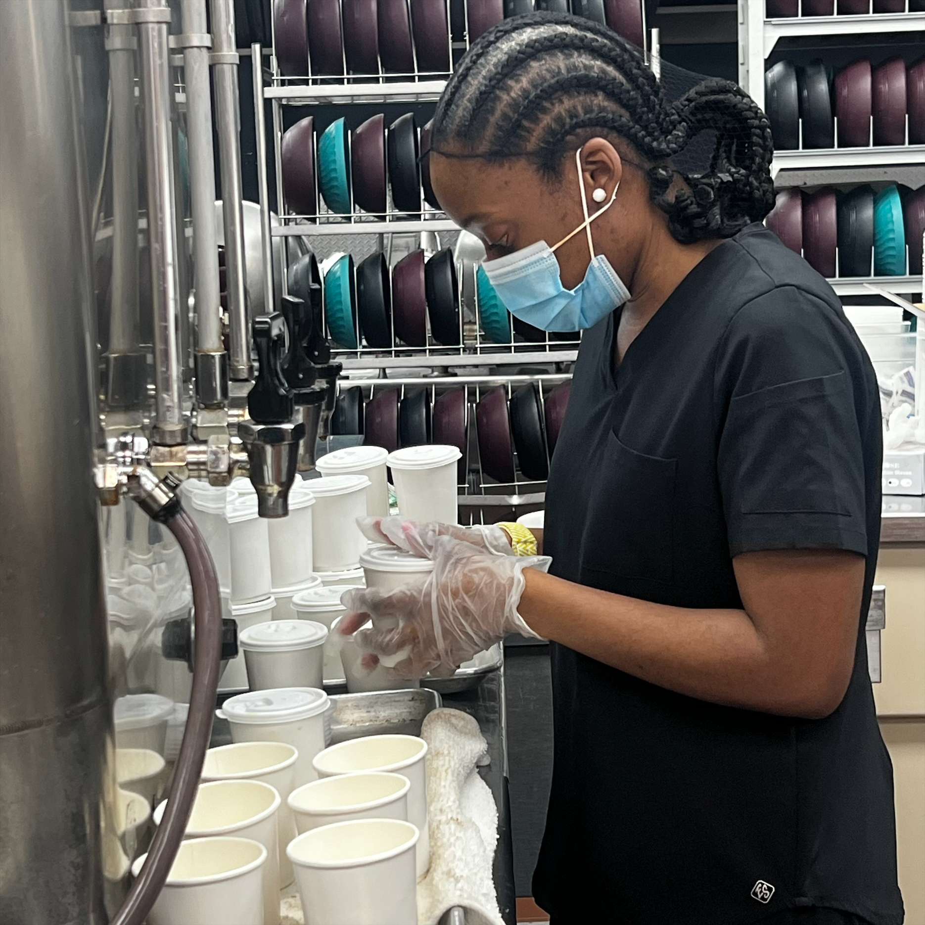 A worker in a black uniform and protective gloves fills cups with a soft serve machine at an ice cream shop, shelves with colorful ice cream containers in the background.