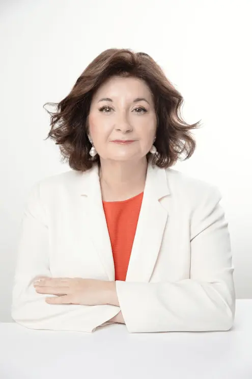 A professional portrait of a woman with shoulder-length brown hair. She wears pearl earrings, a red top, and a white blazer, with her arms crossed in front of her. She has a pleasant expression with a soft smile, and the backdrop is a clean, bright white, enhancing the professional and crisp appearance of the image.