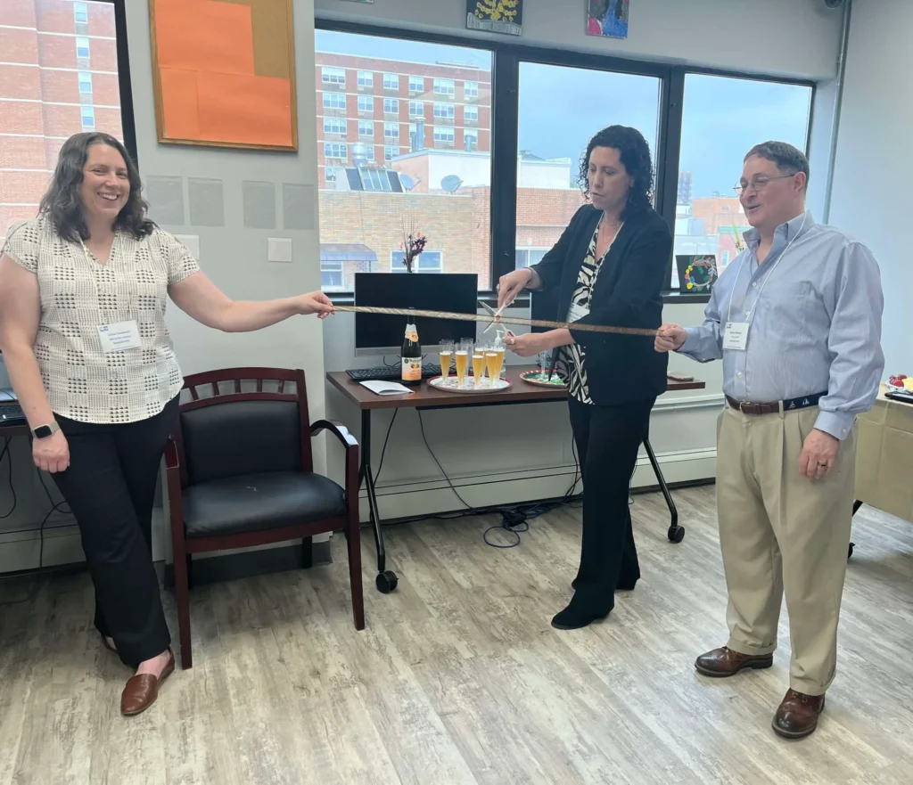 Three people are standing and smiling in an office, holding a ribbon while a woman in the center cuts it with large scissors. There are glasses of champagne and a bottle on a table behind them, indicating a celebratory ribbon-cutting event.