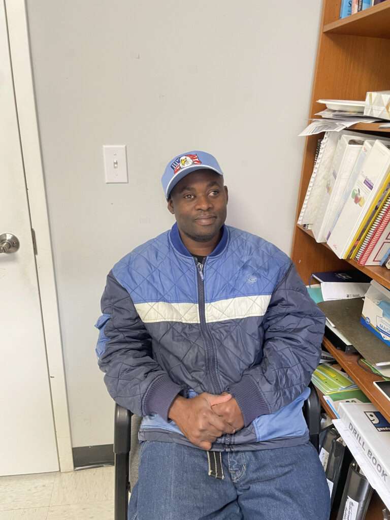 A man is seated in an office chair, wearing a blue quilted jacket and a blue baseball cap with an emblem. He is sitting with his hands clasped together, looking slightly to the side. Behind him is a wooden bookshelf filled with binders and documents, and there is a light switch on the wall to his left. The room appears to be a typical office or workspace.