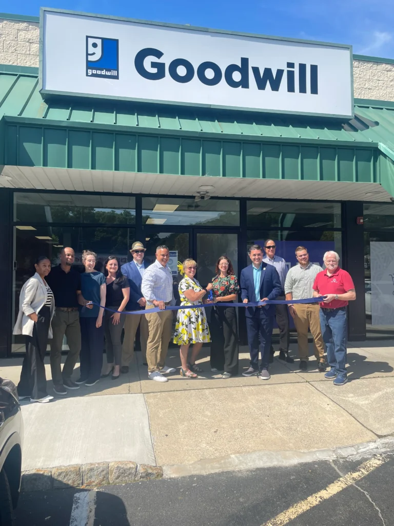 A group of people standing in front of a Goodwill store during a ribbon-cutting ceremony. The group, consisting of both men and women dressed in business casual attire, is holding a blue ribbon. The Goodwill sign is prominently displayed above the entrance. The event appears to be taking place on a sunny day, and the store has a green awning over the entrance.