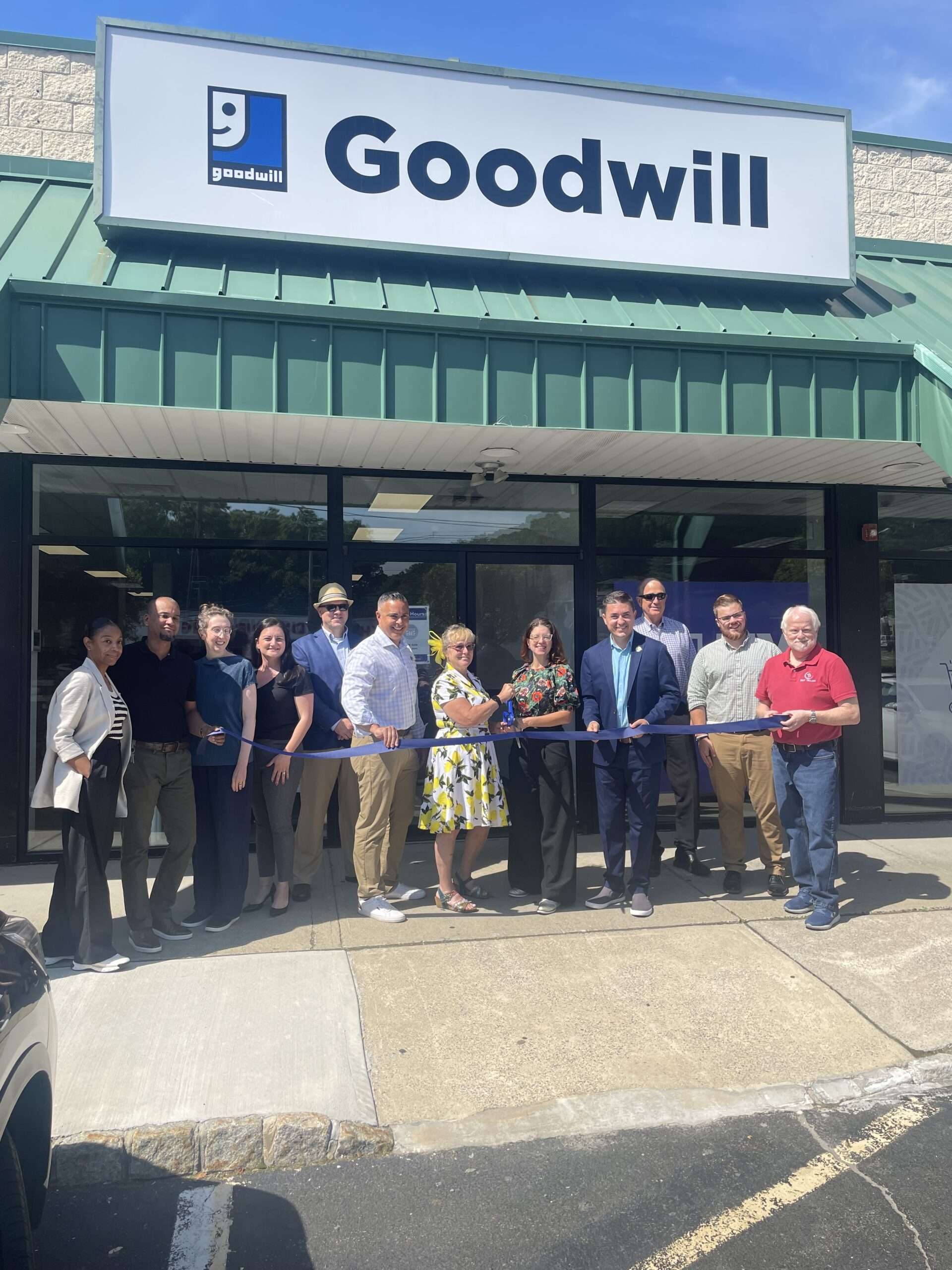A group of people standing in front of a Goodwill store during a ribbon-cutting ceremony. The group, consisting of both men and women dressed in business casual attire, is holding a blue ribbon. The Goodwill sign is prominently displayed above the entrance. The event appears to be taking place on a sunny day, and the store has a green awning over the entrance.