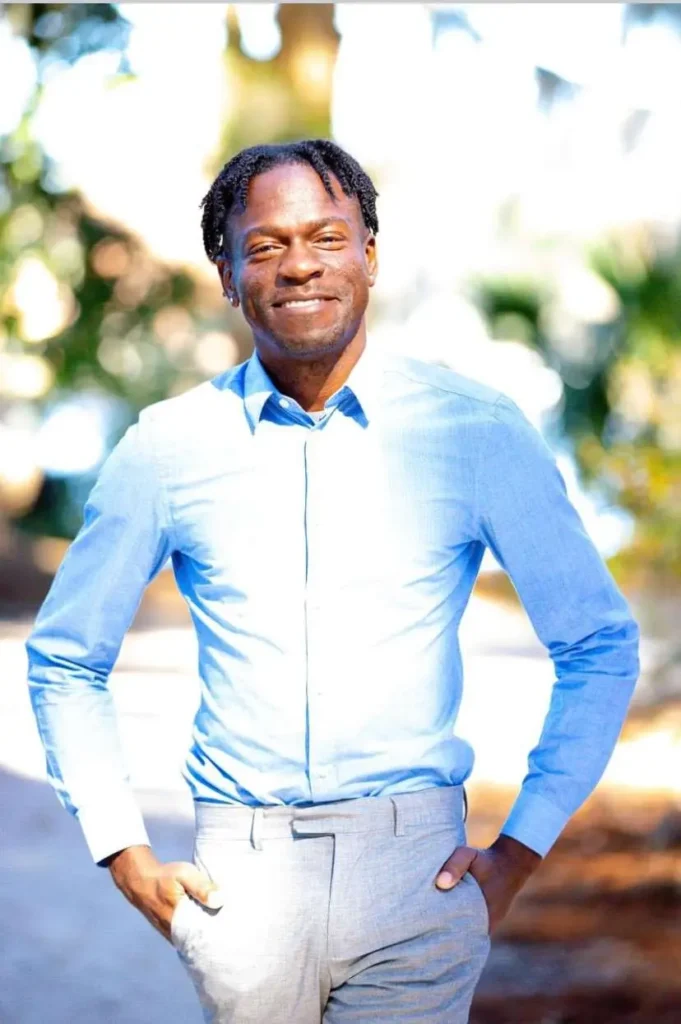 A man in a light blue dress shirt and gray pants stands outdoors, smiling with his hands in his pockets. The background is blurred with hints of greenery and sunlight filtering through trees.