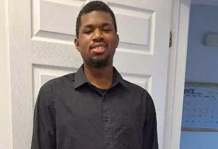 A man stands in front of a white door, wearing a black dress shirt, black pants, and black shoes. He is smiling and appears to be posing for the camera. The background includes a glimpse of a calendar on the wall.