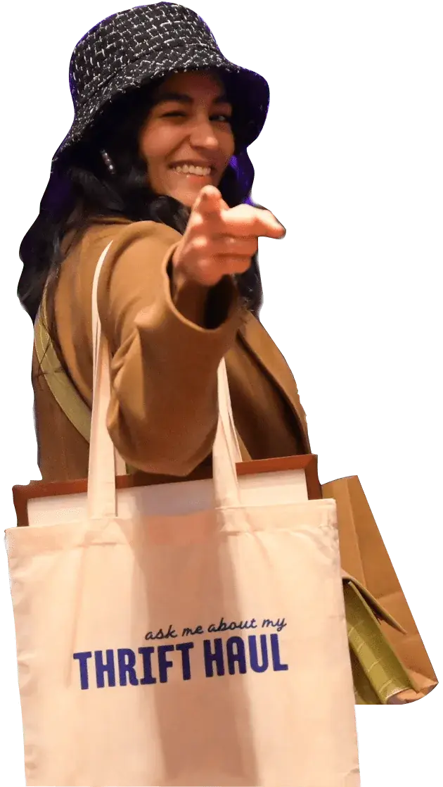 a smiling person wearing a dark textured bucket hat and a brown coat, pointing playfully towards the camera. They are carrying a beige tote bag with the text "ask me about my THRIFT HAUL" printed on it, along with other bags.